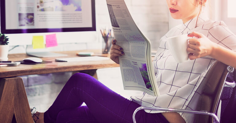 Mulher lendo um informativo com uma xícara de café em mãos e sentada em uma cadeira confortável, se informando sobre o grupo Americas.