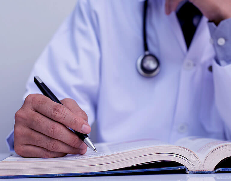 A imagem mostra um médico estudando em um livro e segurando uma caneca.